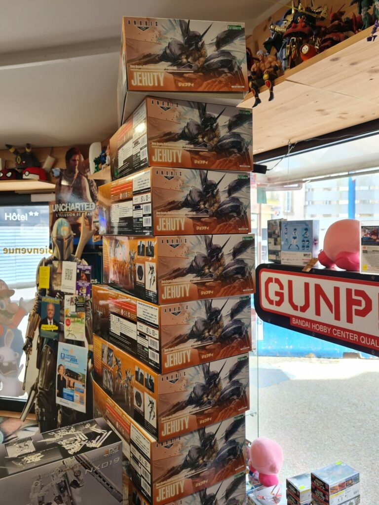 Photographie d'une haute pile verticale de boîtes de maquettes Kotobukiya "Jehuty" tirées du jeu Anubis / Zone of the Enders, exposées dans une boutique spécialisée avec un panneau "GUNPLA" visible à l'arrière-plan.