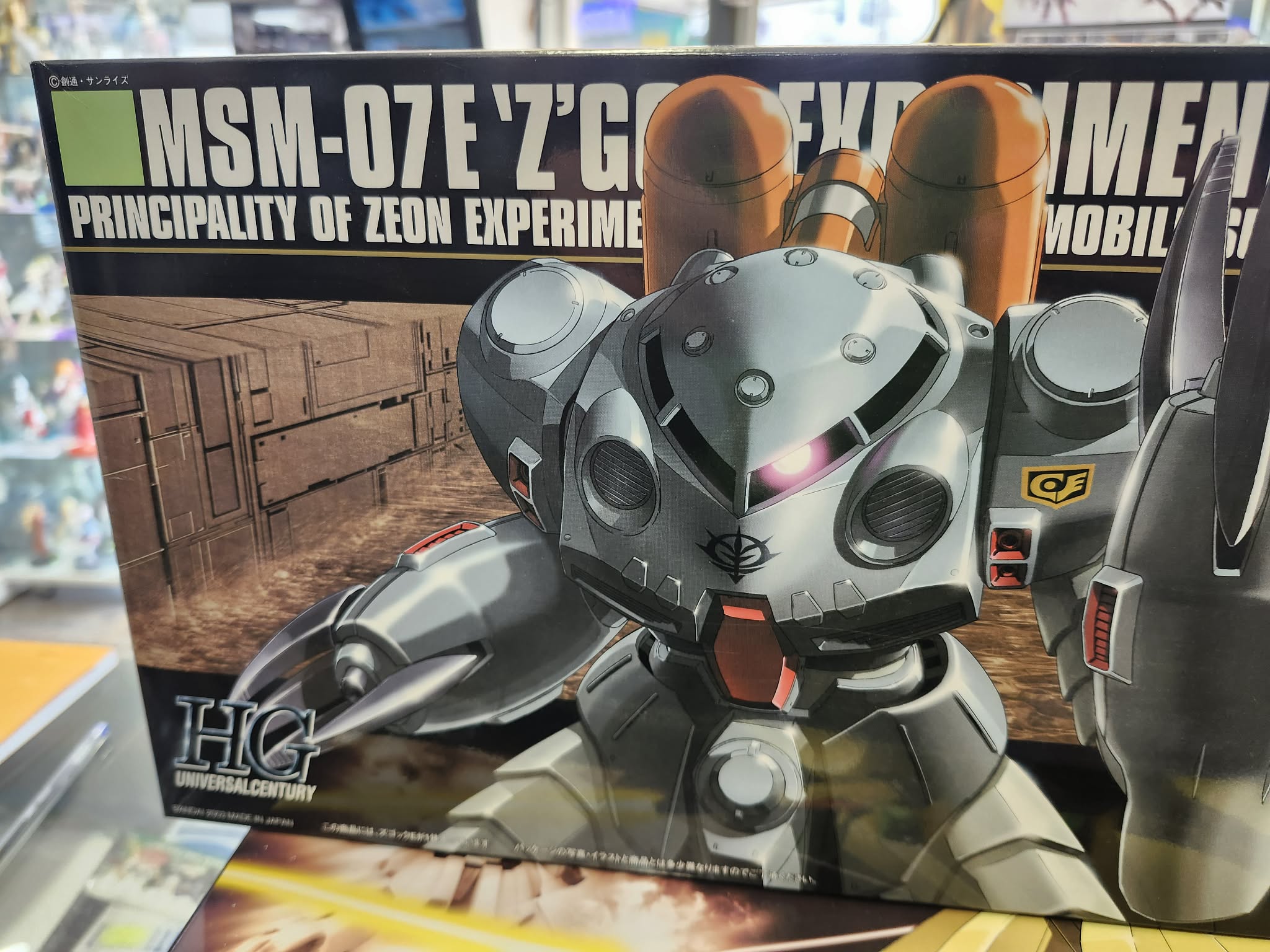 Photographie de la boîte de maquette Bandai Namco High Grade (HG) représentant le mobile suit amphibie expérimental MSM-07E Z'Gok de la Principauté de Zeon, avec un œil rose lumineux et des pinces caractéristiques.