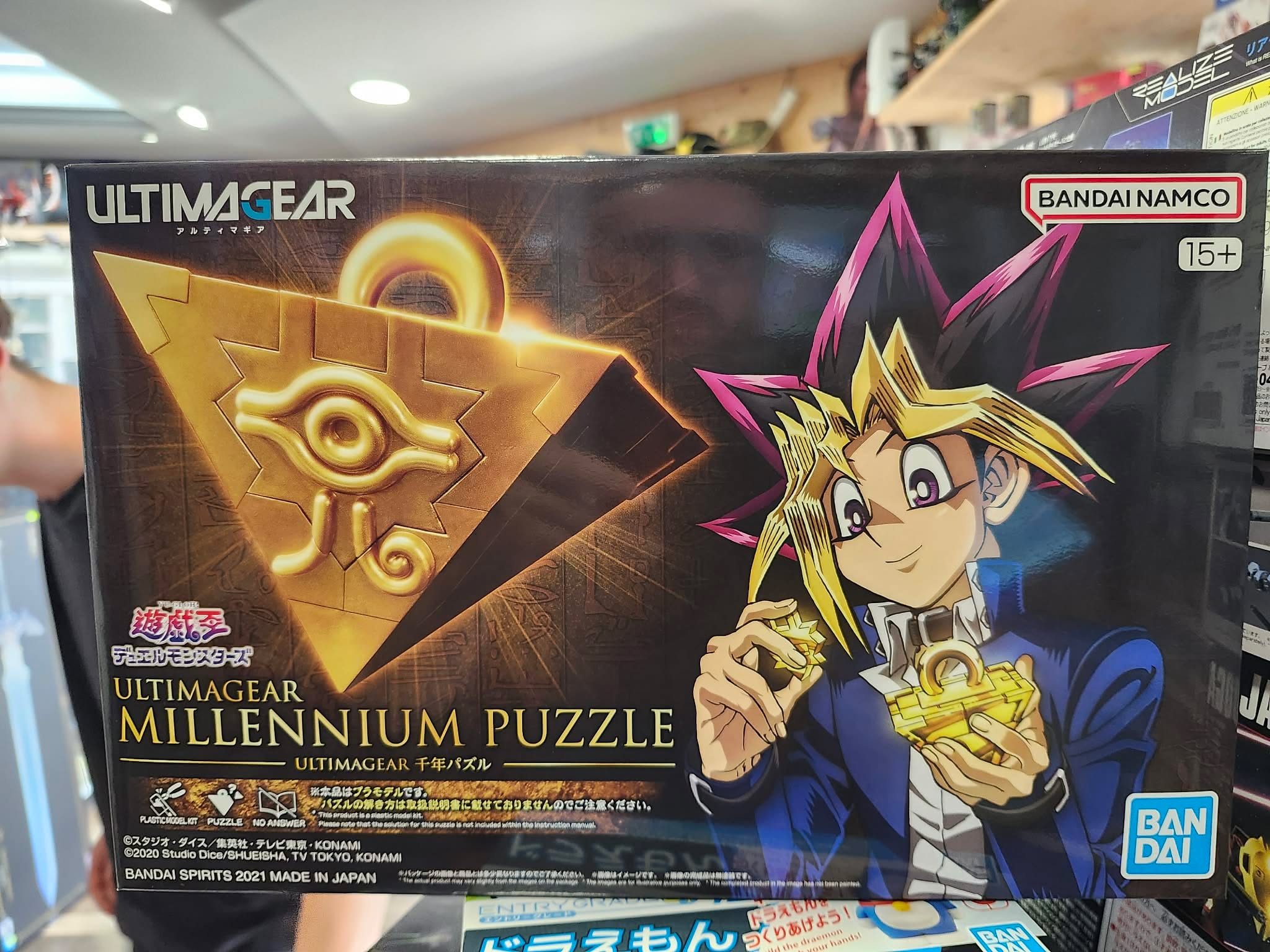 Photographie de la boîte de maquette Bandai Namco de la gamme Ultimagear représentant le Puzzle Millennium (Puzzle du Millénaire) de Yu-Gi-Oh! avec une illustration de Yugi Muto tenant l'objet, prise dans un magasin.